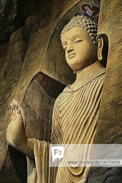 Stehende Buddhastatue in Mönchskleidung  die in eine Kalksteinhöhle gemeißelt und von Licht beleuchtet ist. Rechte Hand in Adhay Mudra Geste. Religion  Tradition  Gebet  Meditation