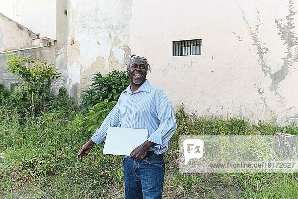 Happy senior architect standing with laptop at site