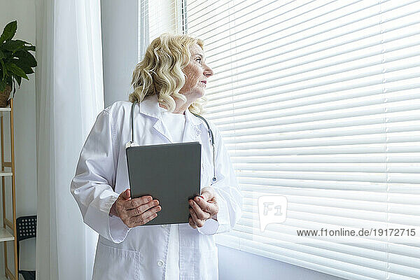 Thoughtful senior doctor with tablet PC looking through window