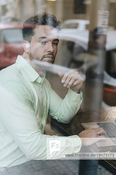 Thoughtful businessman sitting with laptop seen through glass in cafe