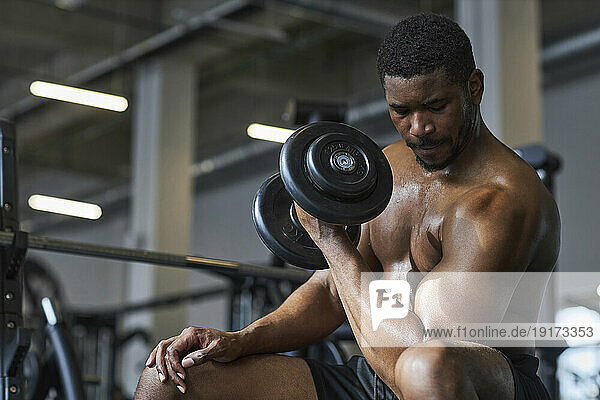 Young shirtless man strengthening biceps with dumbbell in gym
