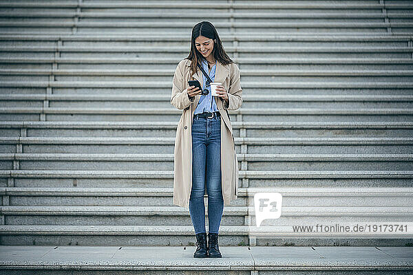 Happy woman with coffee cup using smart phone in front of steps