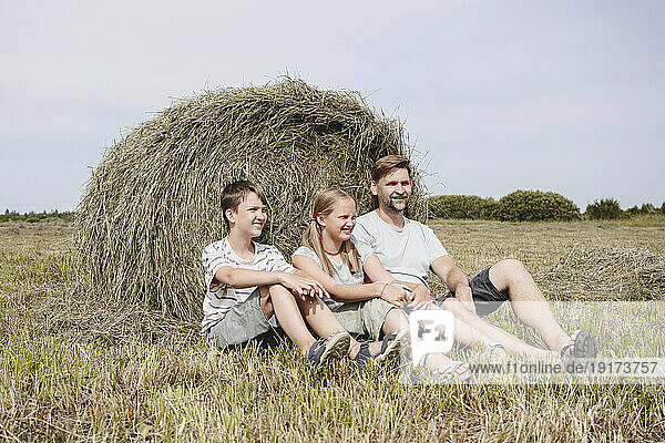 Smiling children sitting with father near hay in field
