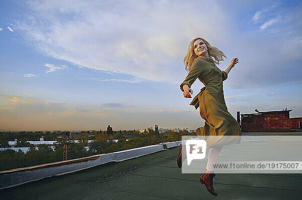 Cheerful blond woman dancing on rooftop under cloudy sky