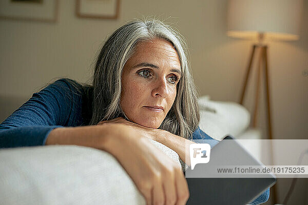 Thoughtful mature woman sitting on sofa with tablet PC