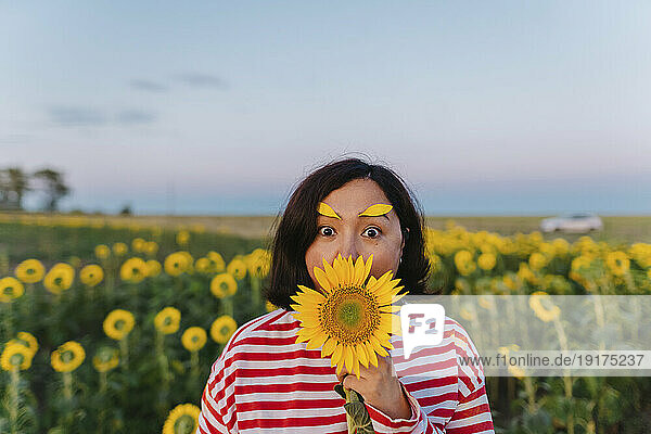 Mature woman with petals on eyebrows covering mouth with sunflower