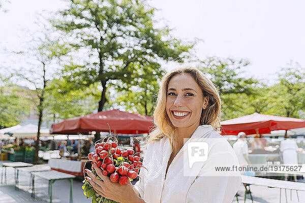 Happy blond woman holding radish in farmer's market
