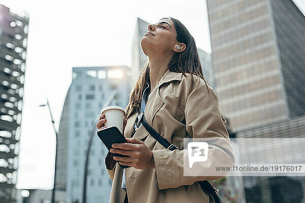 Young woman with eyes closed holding coffee cup and smart phone in city