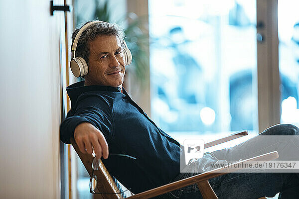 Smiling mature man wearing wireless headphones sitting in armchair
