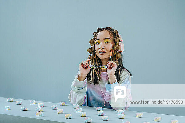 Young woman wearing eyeglasses by marshmallows on table against blue background