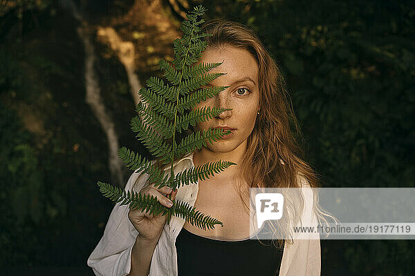 Young blond woman covering face with fern stem