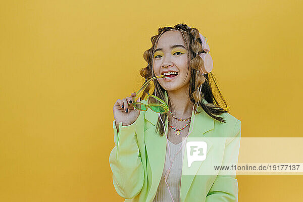 Happy woman with headphones holding sunglasses against yellow background