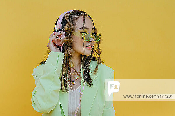 Young woman with sunglasses listening music on headphones against yellow background