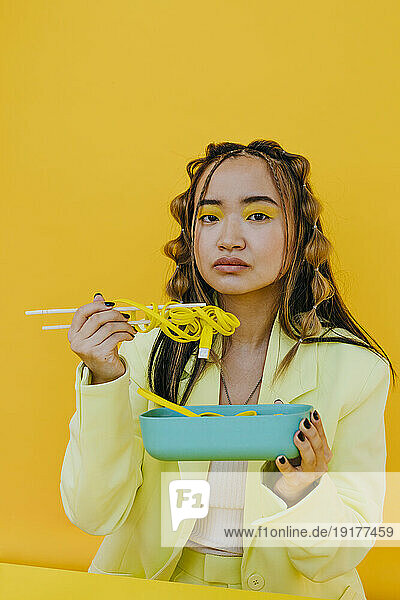 Young woman having wired noodles from bento box against yellow background