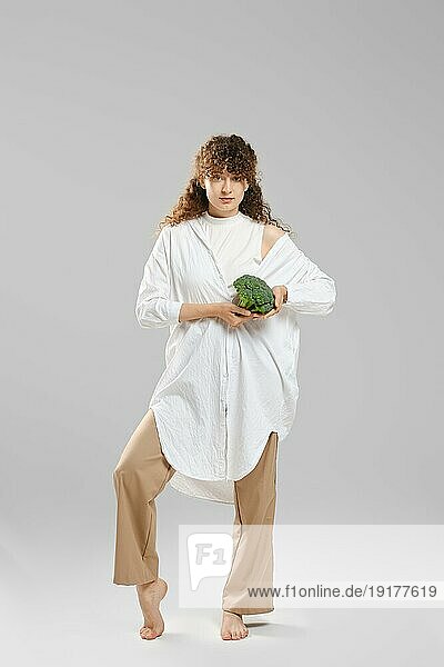 Barfüßige  schlanke Frau in weiten Hosen und Hemd demonstriert Brokkoli
