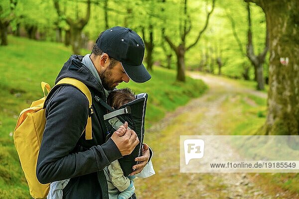 Ein junger Vater mit einem gelben Rucksack geht mit seinem neugeborenen Kind im Rucksack auf einem Waldweg spazieren
