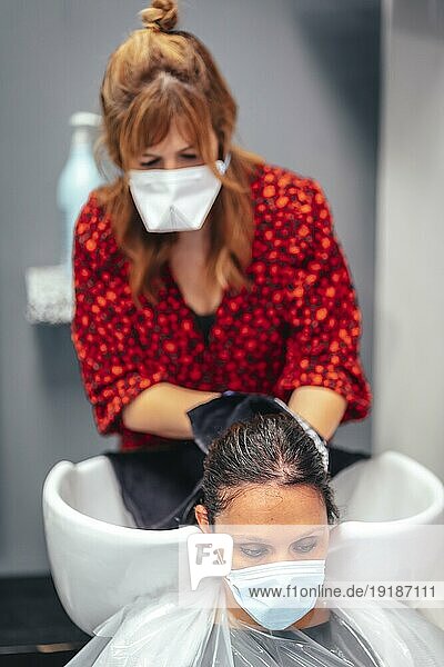 Friseur mit Maske und Handschuhen beim Waschen der Haare eines Kunden mit Wasser und Seife. Wiedereröffnung mit Sicherheitsmaßnahmen für Friseure im Rahmen der Covid 19 Pandemie