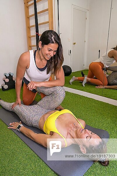 Lateinisches Mädchen beim Sport in einer Sitzung der physikalischen Therapie und Sportosteopathie  Ausbilderin beim Dehnen mit dem Kunden neben einem Spiegel lächelnd  vertikales Foto