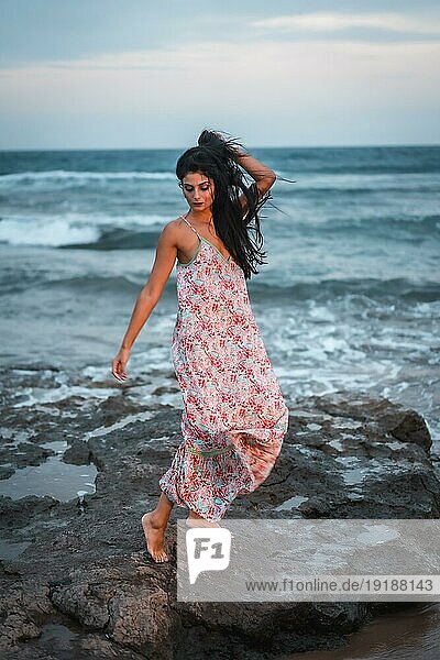 Posing einer brünetten Kaukasierin in einem weißen Stoffkleid mit Blumen  am Strand  die den Urlaub am Meer genießt