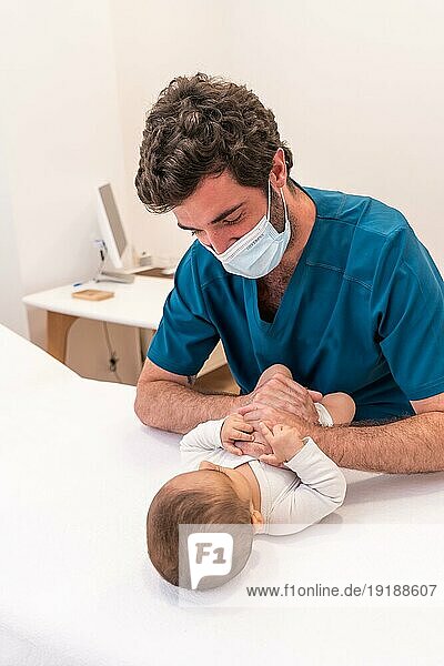 Frontalaufnahme eines jungen Arztes mit Gesichtsmaske  der ein süßes Neugeborenes in einer Klinik untersucht