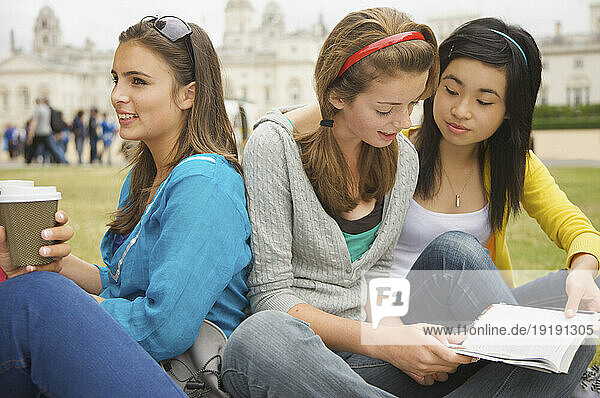 Teenaged girls sitting in front of London Horse Guards Parade drinking coffee and reading a book
