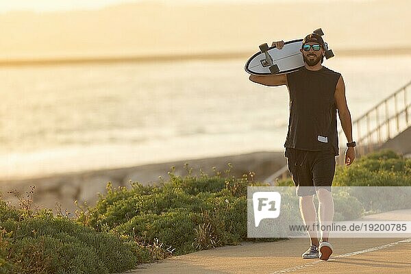Ein Mann geht mit einem Skateboard auf der Straße in Strandnähe spazieren. Mittlere Einstellung