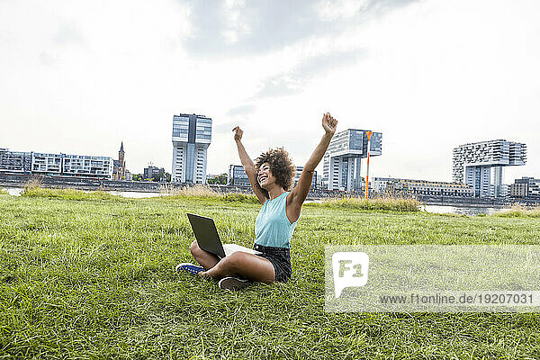 Germany  Cologne  cheering woman sitting on a meadow using laptop