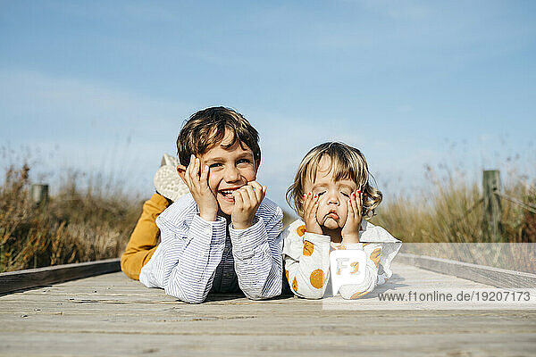 Portrait of boy and his little sister lying side by side on boardwalk pulling funny faces