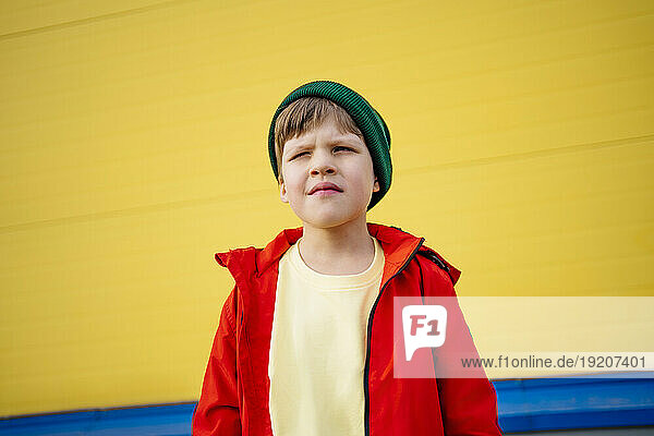 Boy wearing red jacket and knit hat standing in front of yellow wall