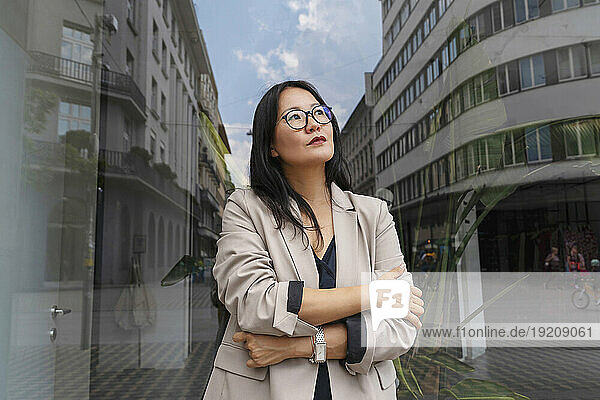 Thoughtful businesswoman with arms crossed day dreaming in front of glass wall