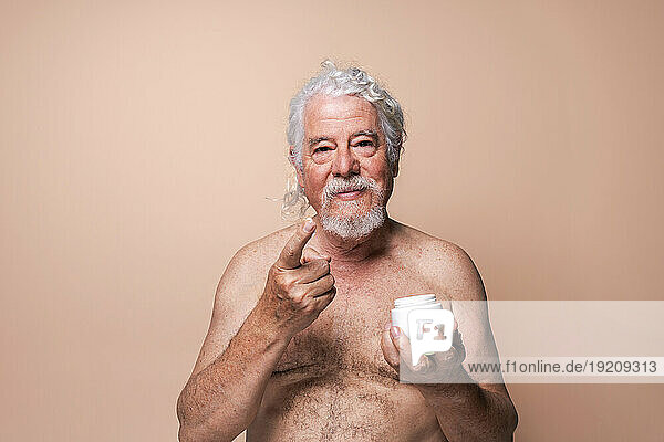 Shirtless senior man holding moisturizer against beige background