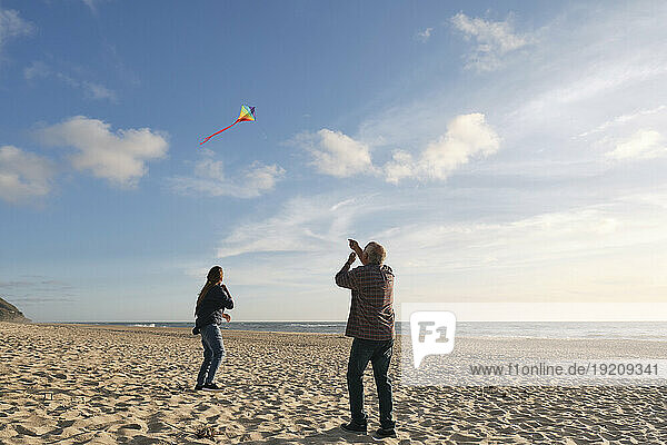 Senior man and woman flying kite at beach on sunny day under cloudy sky