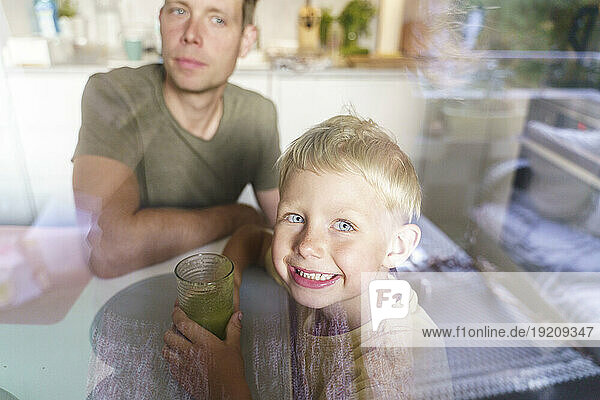 Smiling son holding glass of green juice with father in background