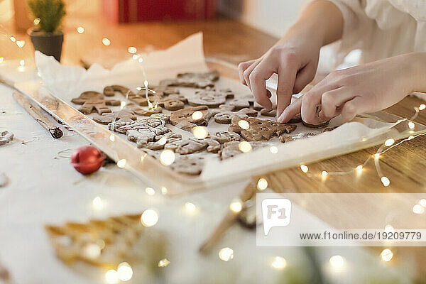 Woman making gingerbread cookies near string lights on table at home