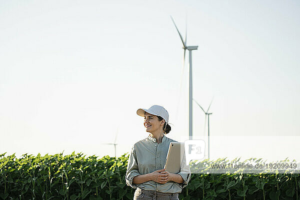 Smiling agronomist holding laptop at field with wind turbines in background