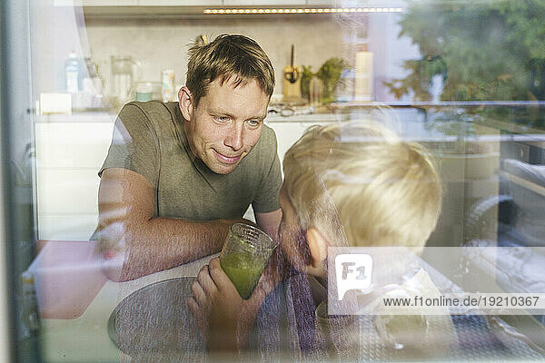 Smiling father looking at son holding glass of green juice in kitchen