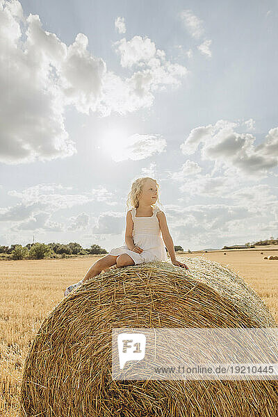 Contemplative blond girl sitting on bale of straw in field on sunny day