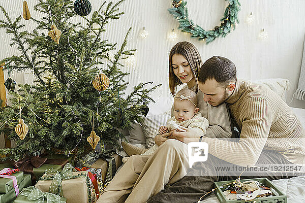 Young parents giving Christmas ornament to baby daughter on bed at home