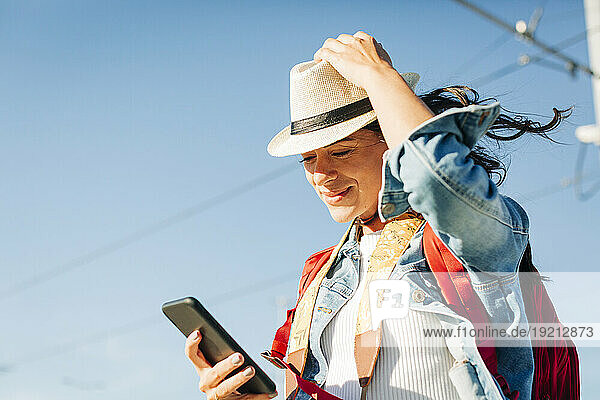 Smiling young woman wearing hat using smart phone under sky