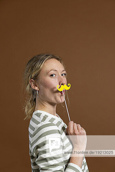 Blond woman holding mustache prop by brown background
