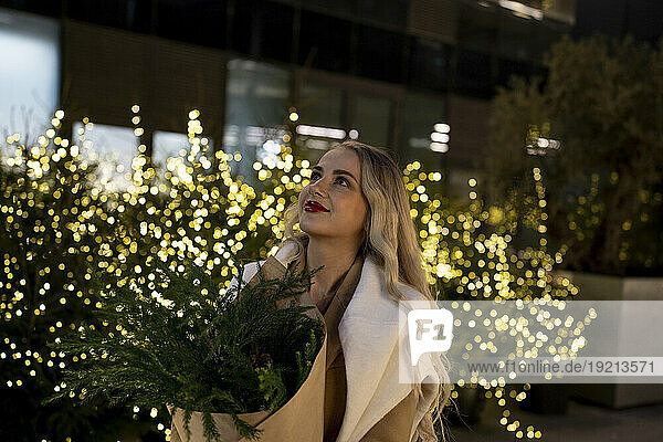 Smiling woman holding bouquet of plants near Christmas lights