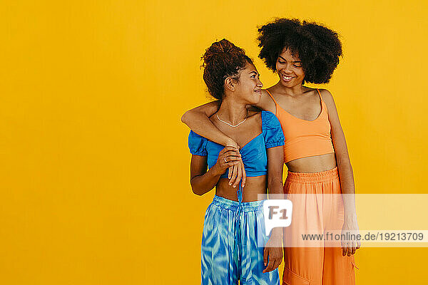 Smiling woman with arm around friend standing against yellow background