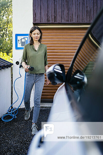 Smiling woman holding electric plug near car at station
