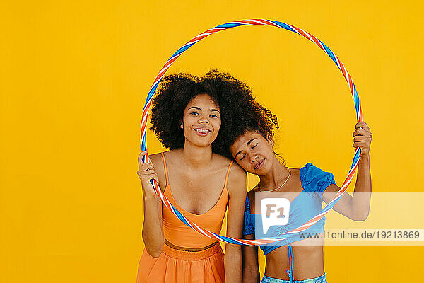 Woman leaning on friend holding plastic hoop against yellow background