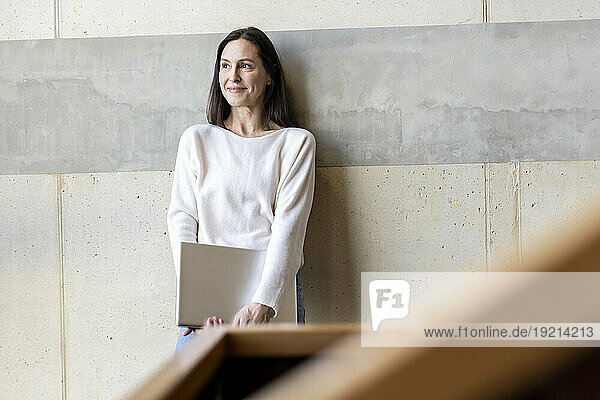 Thoughtful mature businesswoman standing with laptop in front of wall