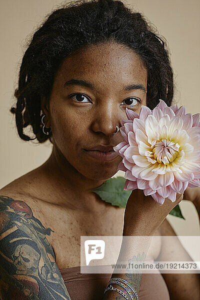 Young woman with tattoo and acne scars holding flower against brown background