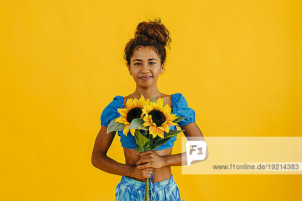 Smiling young woman holding sunflowers standing against yellow background