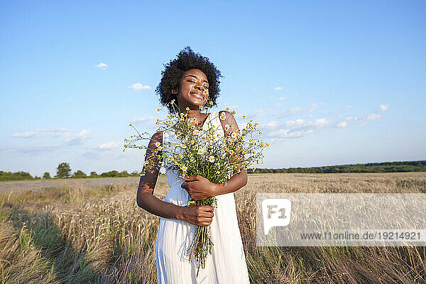 Smiling young woman holding bunch of daises in field under sky