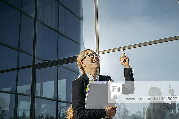 Cheerful businesswoman pointing up and standing by glass building