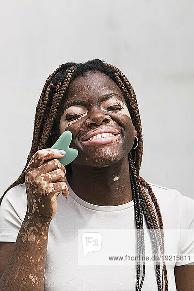 Smiling young woman using gua sha in front of white wall